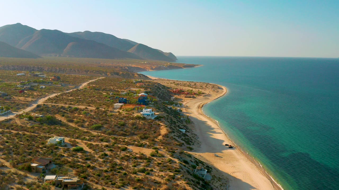 Aerial frame over El Sargento La Ventana, Mexico, with a pale sand curve meeting turquoise water, dry scrub lots and homes in the foreground, mountain ridges in back, and no visible people.