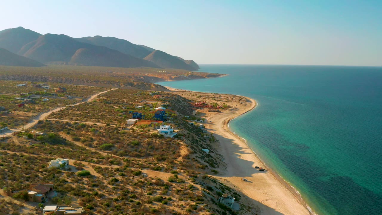 Aerial coastline frame from El Sargento La Ventana, Mexico, with turquoise sea filling the right side, scrubland and scattered homes on the left, dark mountains behind, and no visible people.