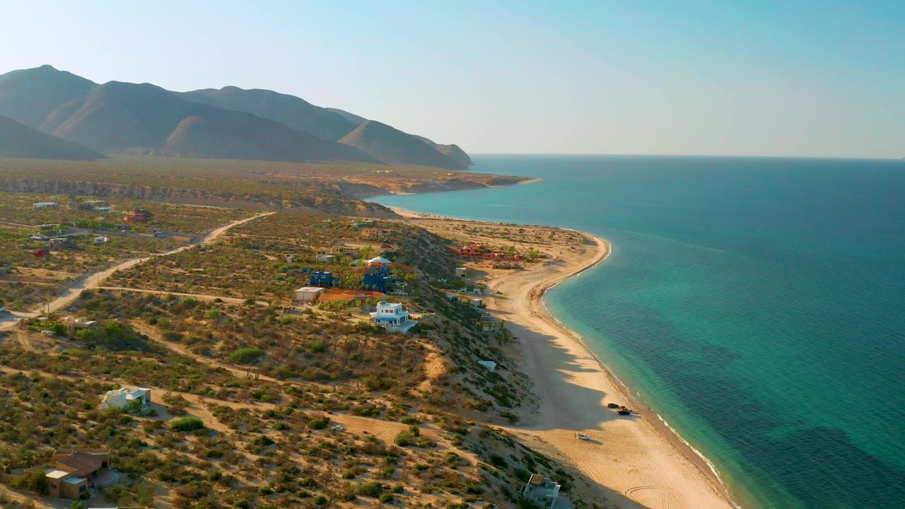 High coastal view of beach, blue water, dry scrub, dirt roads, low houses, and mountains.