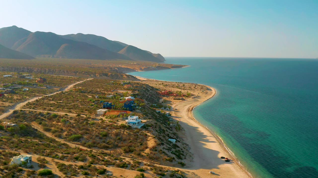 High aerial view in El Sargento La Ventana, Mexico with the beach arc shifted slightly lower in frame, blue-green water to the right, desert lots and houses to the left, mountains in back, and no visible people.