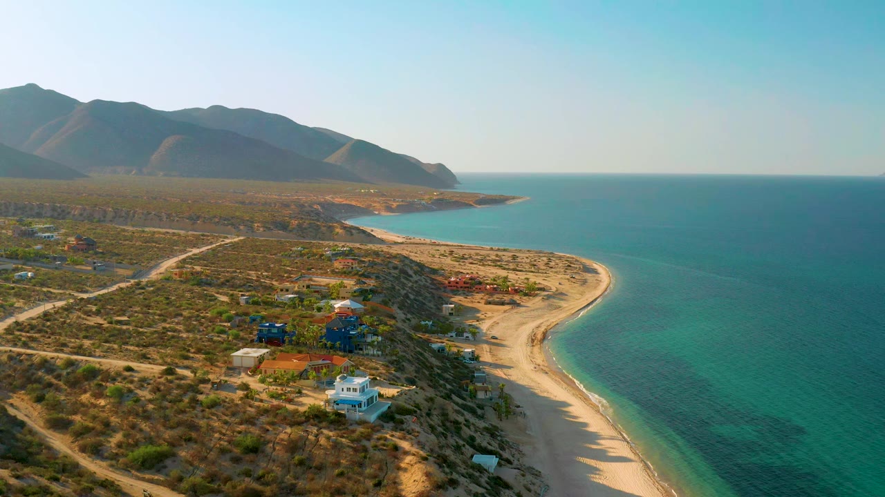 Aerial view in El Sargento La Ventana, Mexico, with white and blue houses in the lower frame, sandy beach along the right edge, turquoise water, distant mountains, and no visible people.