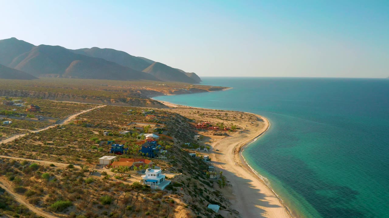 Aerial frame from El Sargento La Ventana, Mexico, showing the shoreline bend more centered, with residential houses, dry terrain, blue-green shallows, mountains, and no visible people.