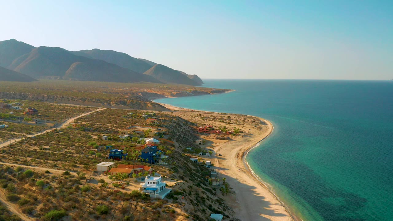 High view of dry coastal lots, scattered homes, sandy shore, blue water, and mountain backdrop.