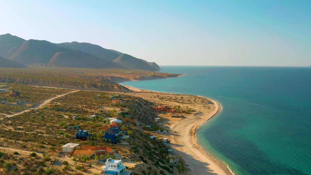 Wide drone view of desert coastline, sandy beach, ocean, and mountain backdrop.
