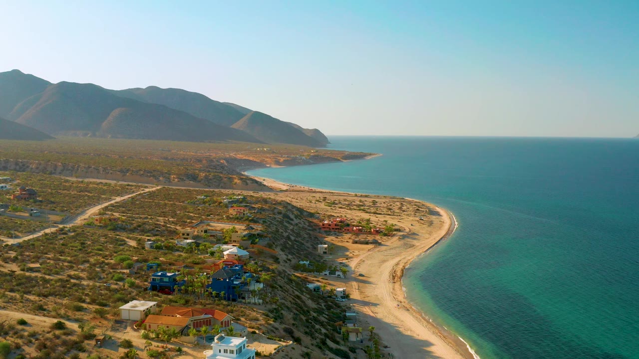 High oblique aerial over El Sargento La Ventana showing the beach arc beside calm turquoise sea, low desert development in the foreground, mountains beyond the shore, and no visible people.