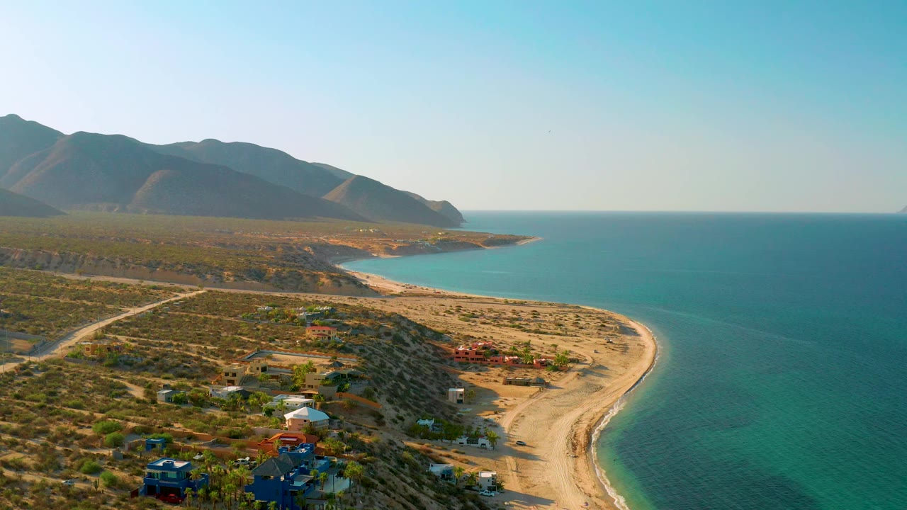 Aerial frame over the El Sargento La Ventana coast showing the sandy beach wrapping around the bay, turquoise water, low desert vegetation, a few buildings, distant mountains, and no visible people.