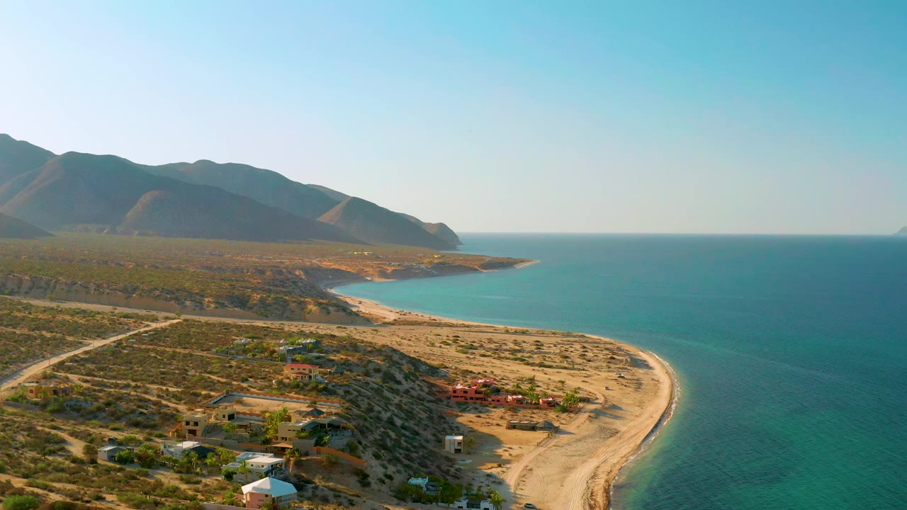 The second frame keeps the El Sargento La Ventana coastline wide, with hillside buildings lower in frame, the sandy beach sweeping right, blue water, and no visible people.