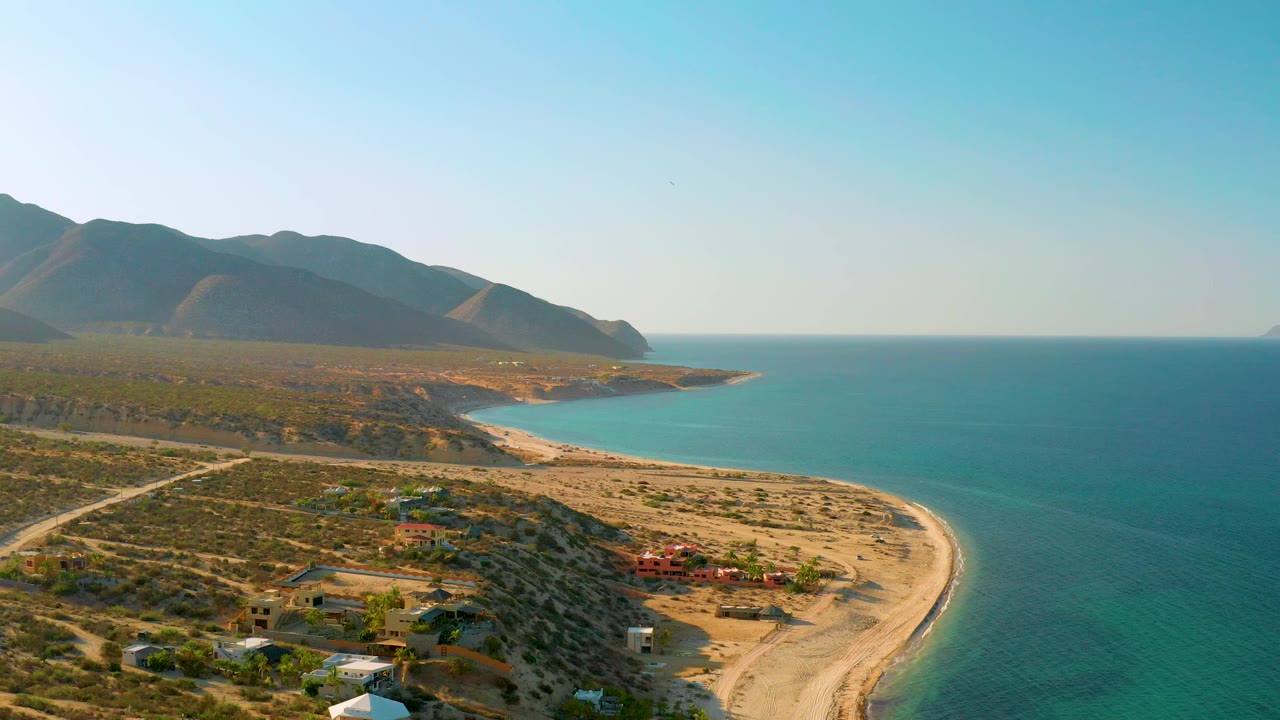 A high view over El Sargento La Ventana shows more open water to the right, a pale beach along the cove, dark mountain slopes on the left, and no visible people.