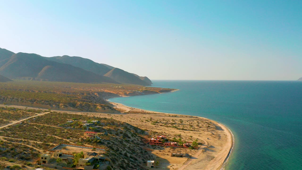Opening aerial view over El Sargento La Ventana, Mexico, with blue-green sea beside a pale sand beach, scattered hillside houses, distant mountains, and no visible people.