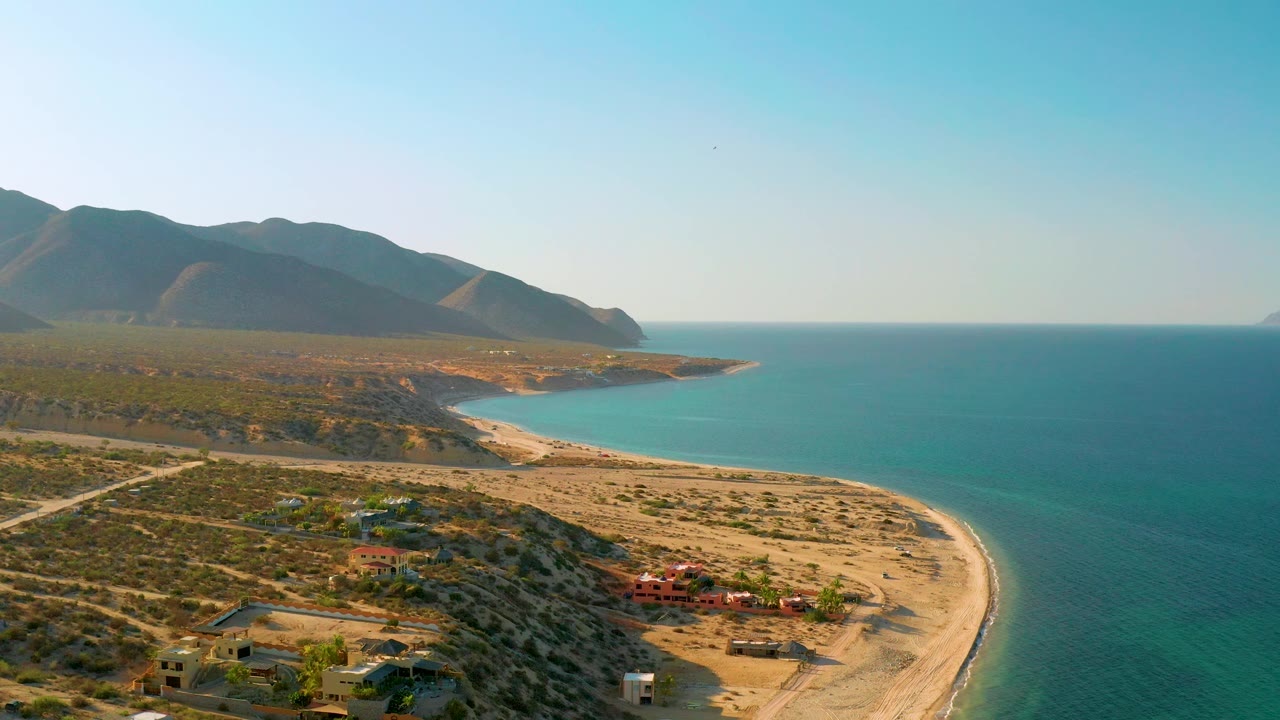 At 0:02, the El Sargento La Ventana coastline remains in a high view, with blue ocean, a narrow pale beach, arid hills, small buildings, and no visible people.