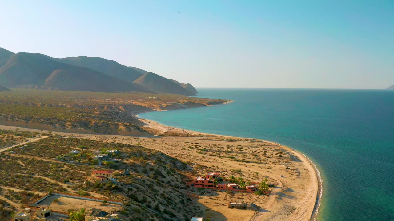 One second into the clip, the aerial view holds on the El Sargento La Ventana shoreline, with the beach arc clearer along the right edge, small buildings below, distant mountains, and no visible people.