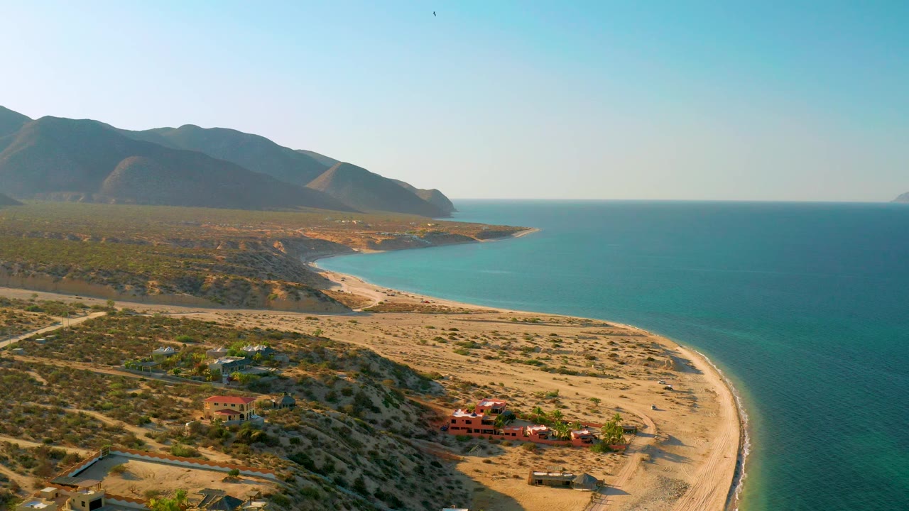 At 0:02, the El Sargento La Ventana coastline remains in a wide aerial composition, with turquoise water filling the right side, arid land and roads on the left, mountains beyond, and no visible people.