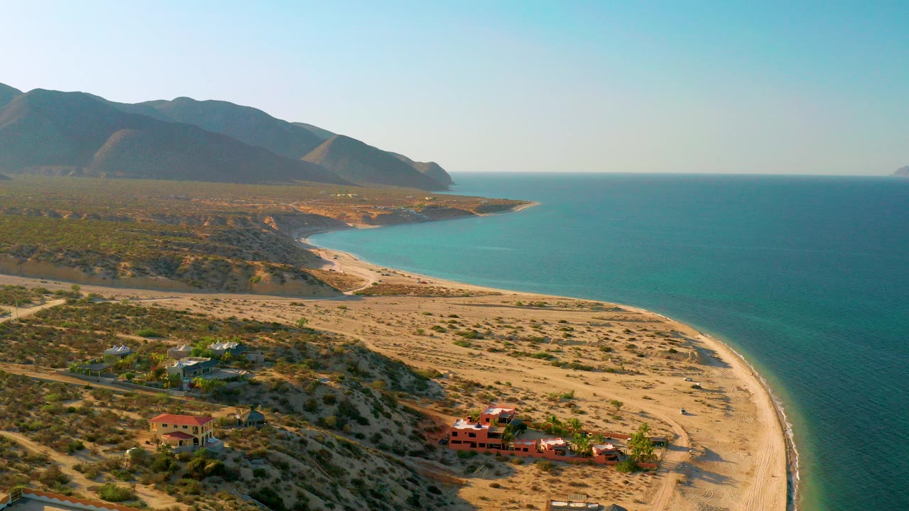 Aerial frame over El Sargento La Ventana, Mexico showing a curved sandy beach, blue sea to the right, scrubland and houses below, mountains in the distance, and no visible people.