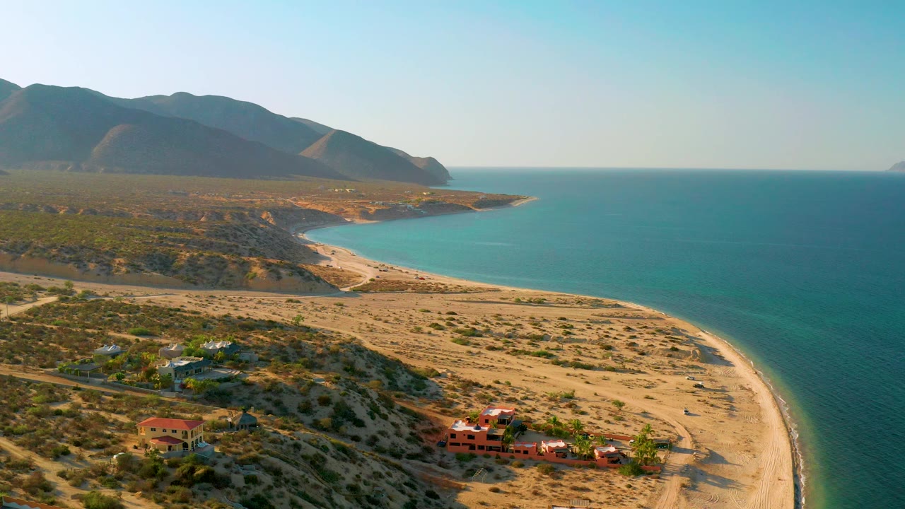 Drone frame over El Sargento La Ventana, Mexico showing the beach arc along the lower right edge, broad blue water, scrub-covered terrain, a small group of houses near the sand, distant mountains, and no visible people.