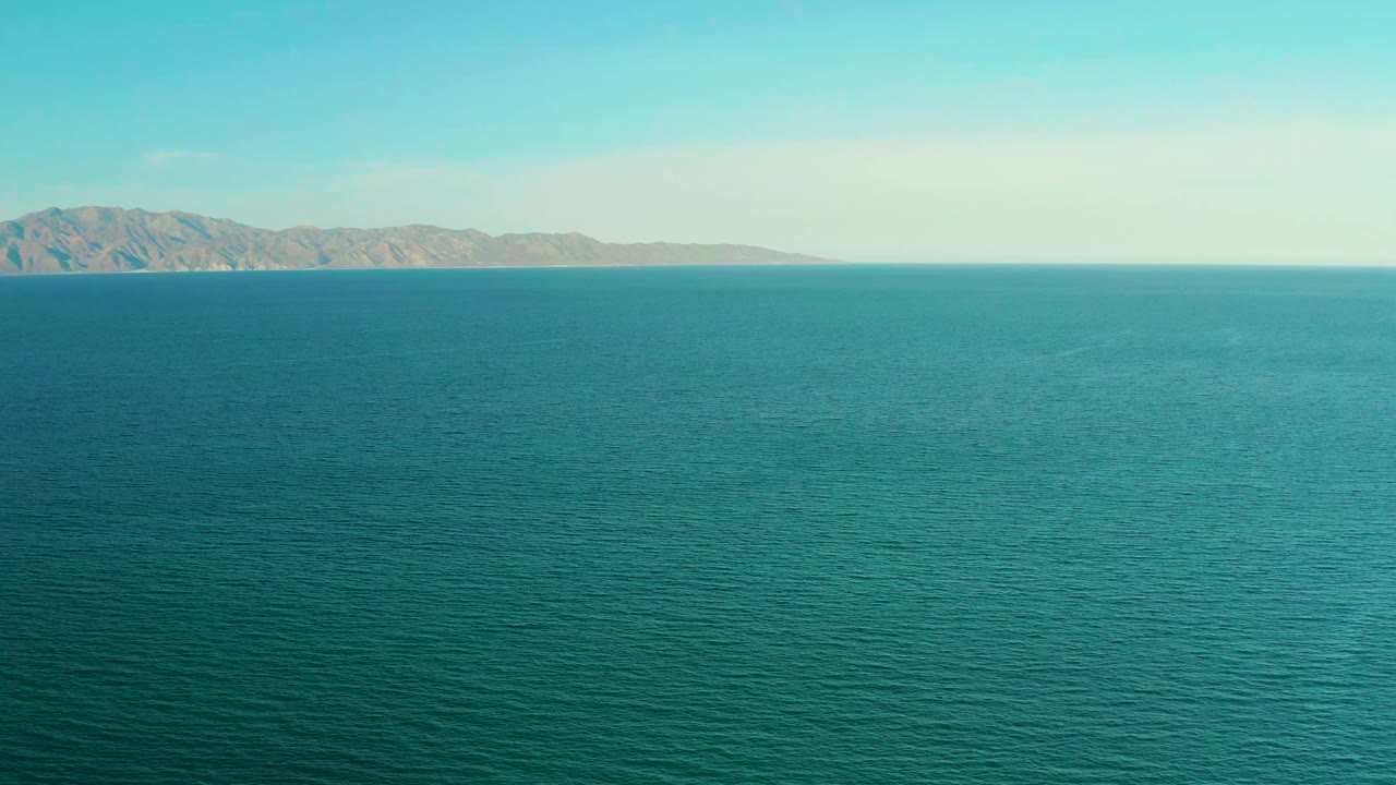 Opening frame of teal ocean water off El Sargento La Ventana, Mexico, with a tan landmass stretching across the distant horizon and no visible people.