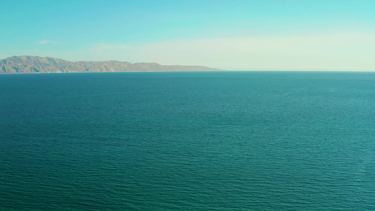 Teal ocean fills the frame under a pale sky, with the far shoreline and low landforms visible near the top edge in El Sargento La Ventana, Mexico, and no visible people.