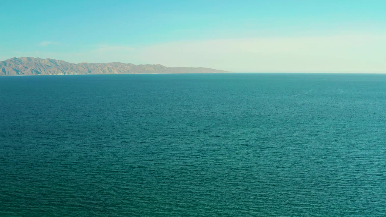 A broad field of teal water appears in the foreground and midground, with distant coastal hills along the left horizon in El Sargento La Ventana, Mexico, and no visible people.