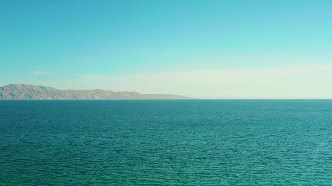 At 0:02, the blue-green ocean shows fine ripple texture below a broad sky, with distant mountains stretching across the left background in El Sargento La Ventana and no visible people.