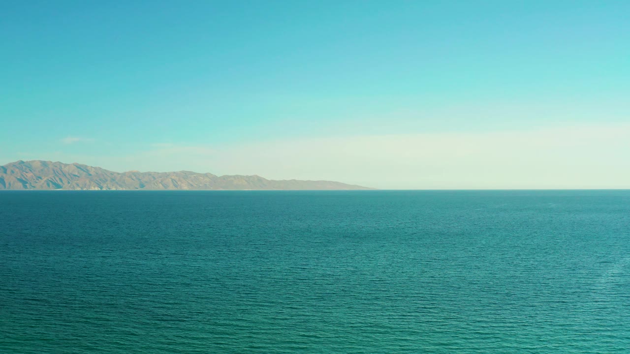 At 0:03, the sampled frame keeps the ocean in the foreground and the El Sargento La Ventana mountain horizon low across the frame, with pale daylight and no visible people.