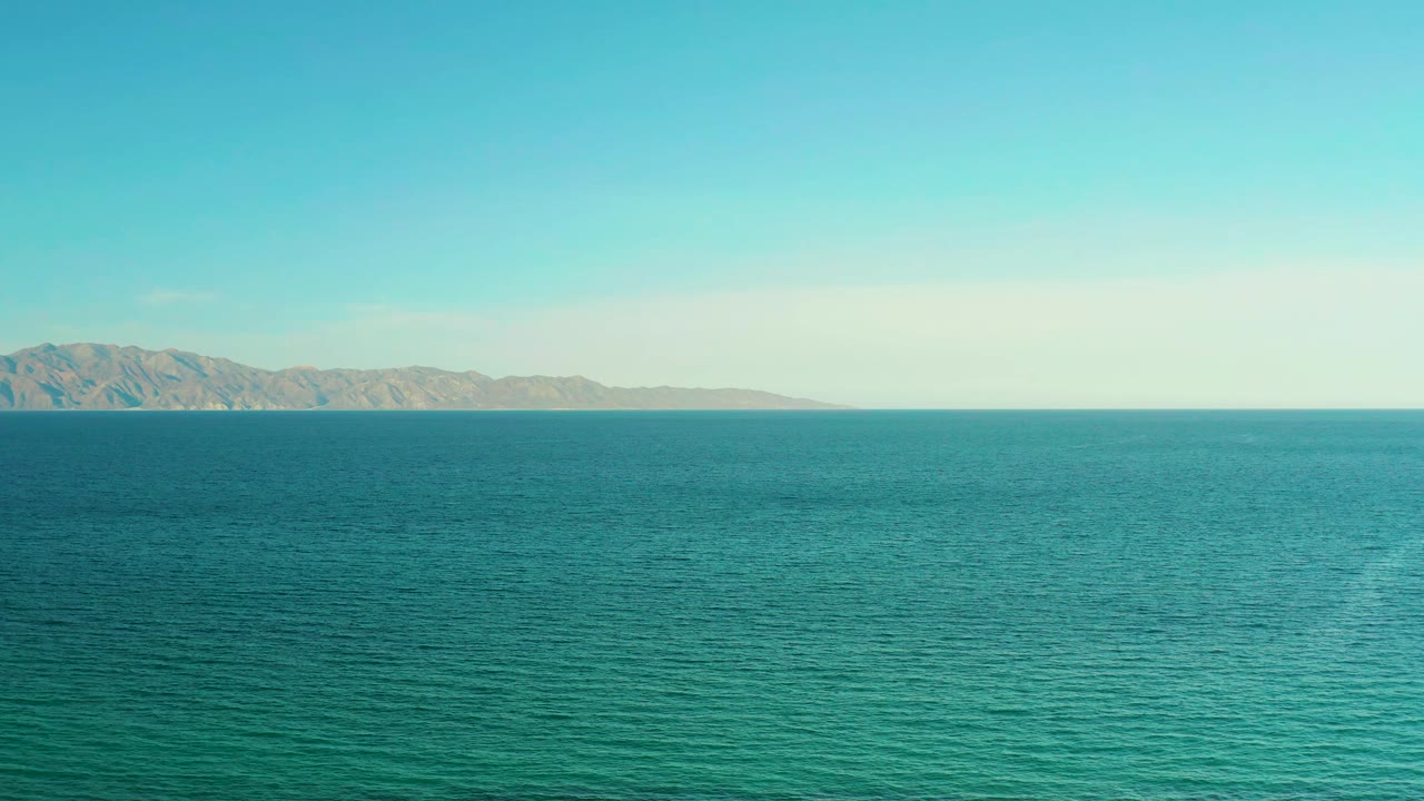 Wide coastal frame from El Sargento La Ventana, Mexico, showing turquoise water in the foreground, a pale sky, and distant mountains with no visible people.