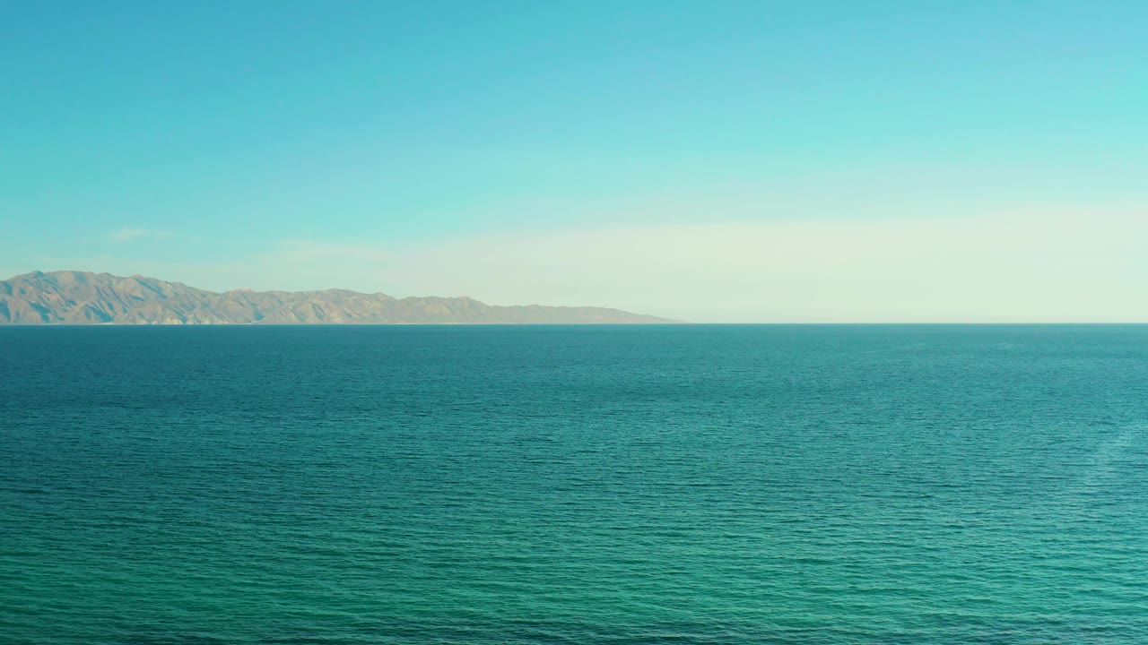 Blue-green sea fills most of the frame while distant mountains sit left of center on the horizon near El Sargento La Ventana, Mexico, with no visible people.