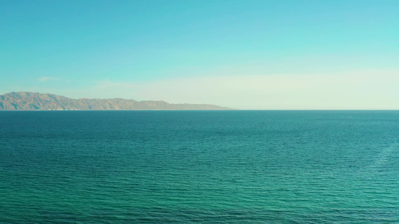 Mid-clip view of the El Sargento La Ventana coastline from offshore or elevated perspective, with rippled blue water and low mountains on the far left, no visible people.