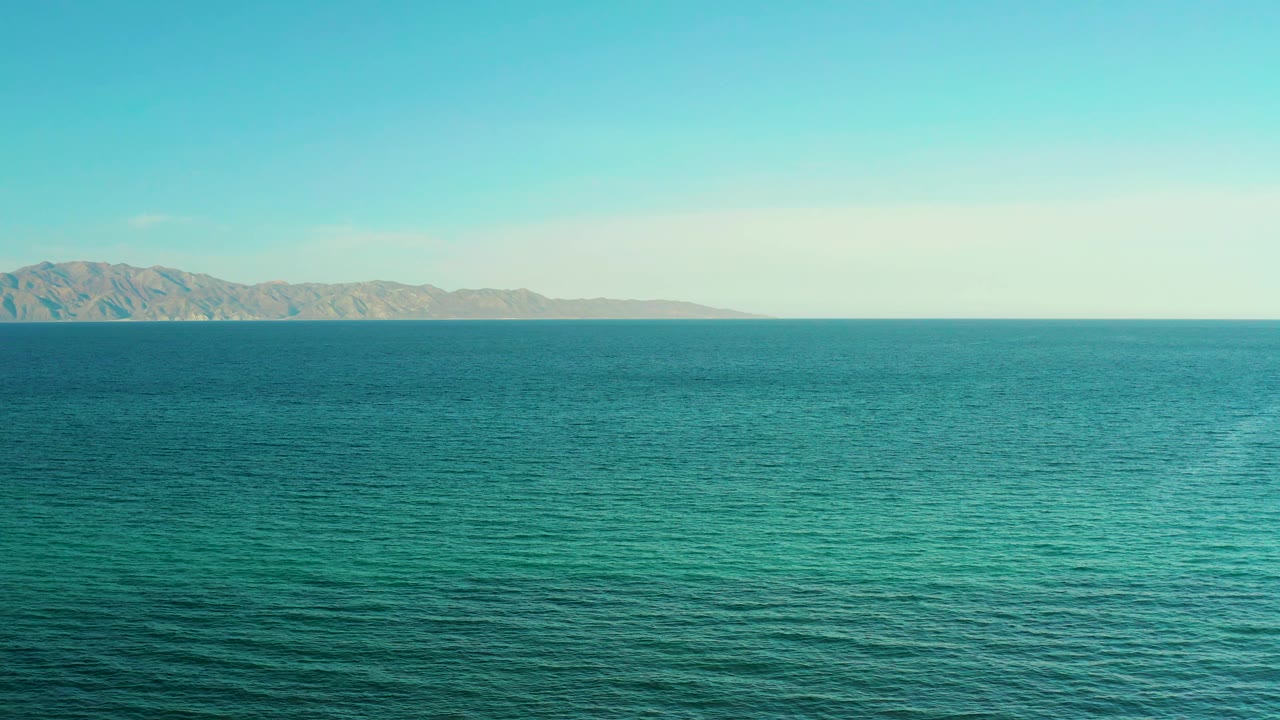 The second frame keeps the ocean dominant, with the mountain ridge slightly left of center, a level horizon, pale blue sky, and no visible people.