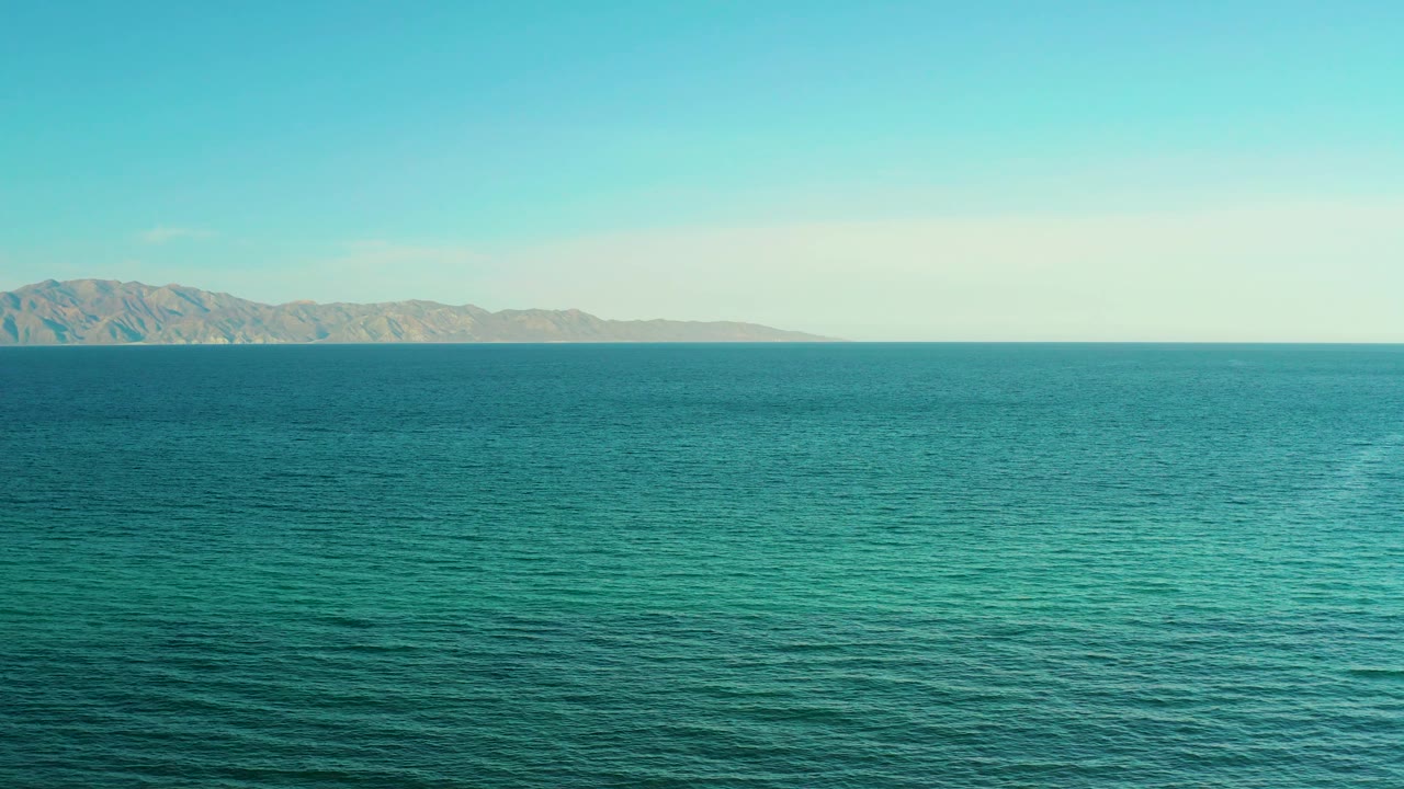 Blue-green water stretches to a distant mountain range at El Sargento La Ventana, Mexico, with light haze near the horizon and no visible people.