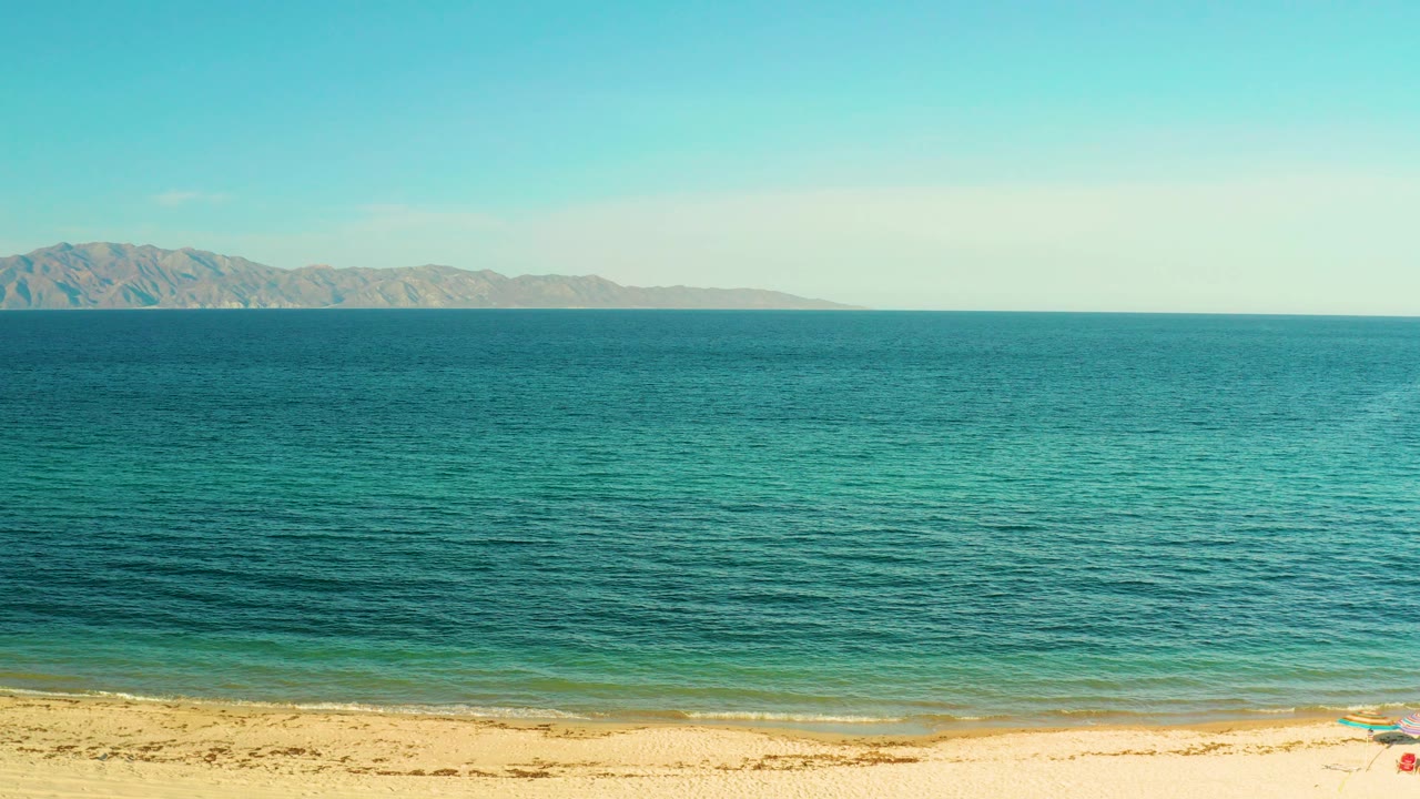Wide elevated view of El Sargento La Ventana beach with sandy shore, turquoise water, distant mountains, pale sky, and no visible people.