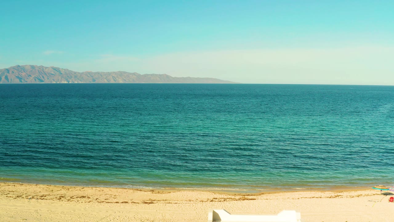 Wide beach frame in El Sargento La Ventana with turquoise shallows, darker offshore water, distant rocky land, open sky, and no visible people.