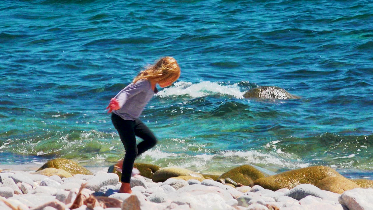 At 0:00, a young blonde girl steps carefully across pale shoreline rocks in El Sargento La Ventana, Mexico, with turquoise water and small breaking waves behind her.