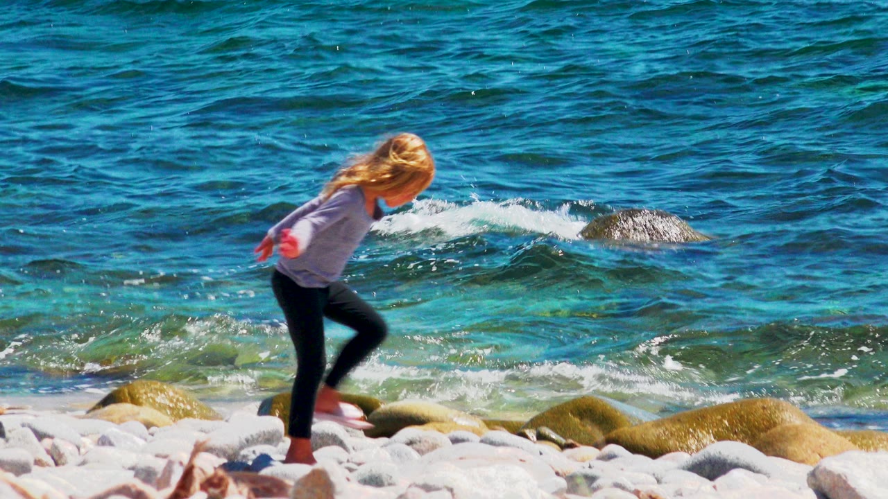 Young girl mid-step on shoreline rocks with turquoise water behind her.