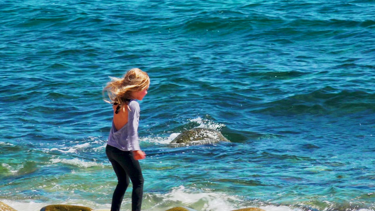 At 0:02, the girl has moved farther right along the shoreline, with her back partly turned and white surf breaking over dark rocks in the blue water behind her in El Sargento La Ventana, Mexico.