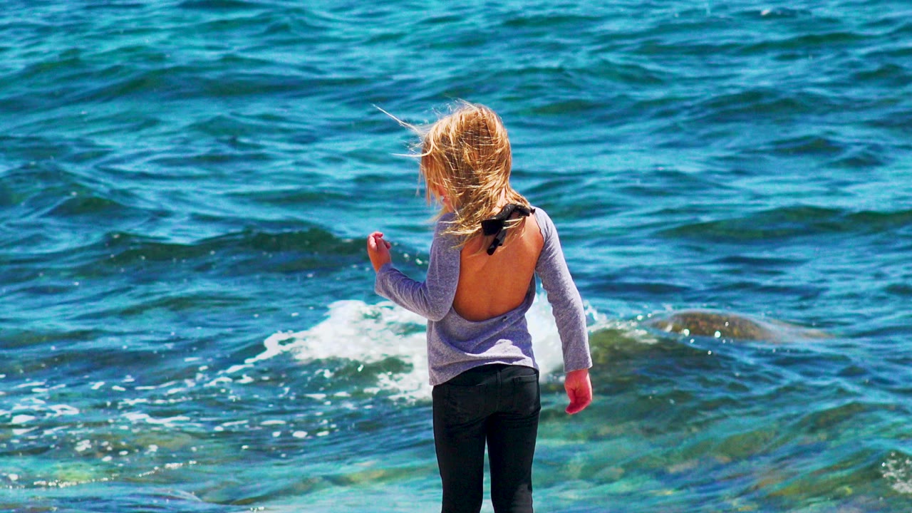 A young girl in a gray top stands with her back partly to camera near blue waves on the rocky shoreline in El Sargento La Ventana, Mexico.
