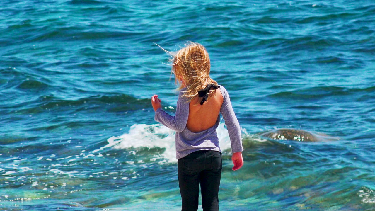 Young girl facing the ocean as waves break over rocks.