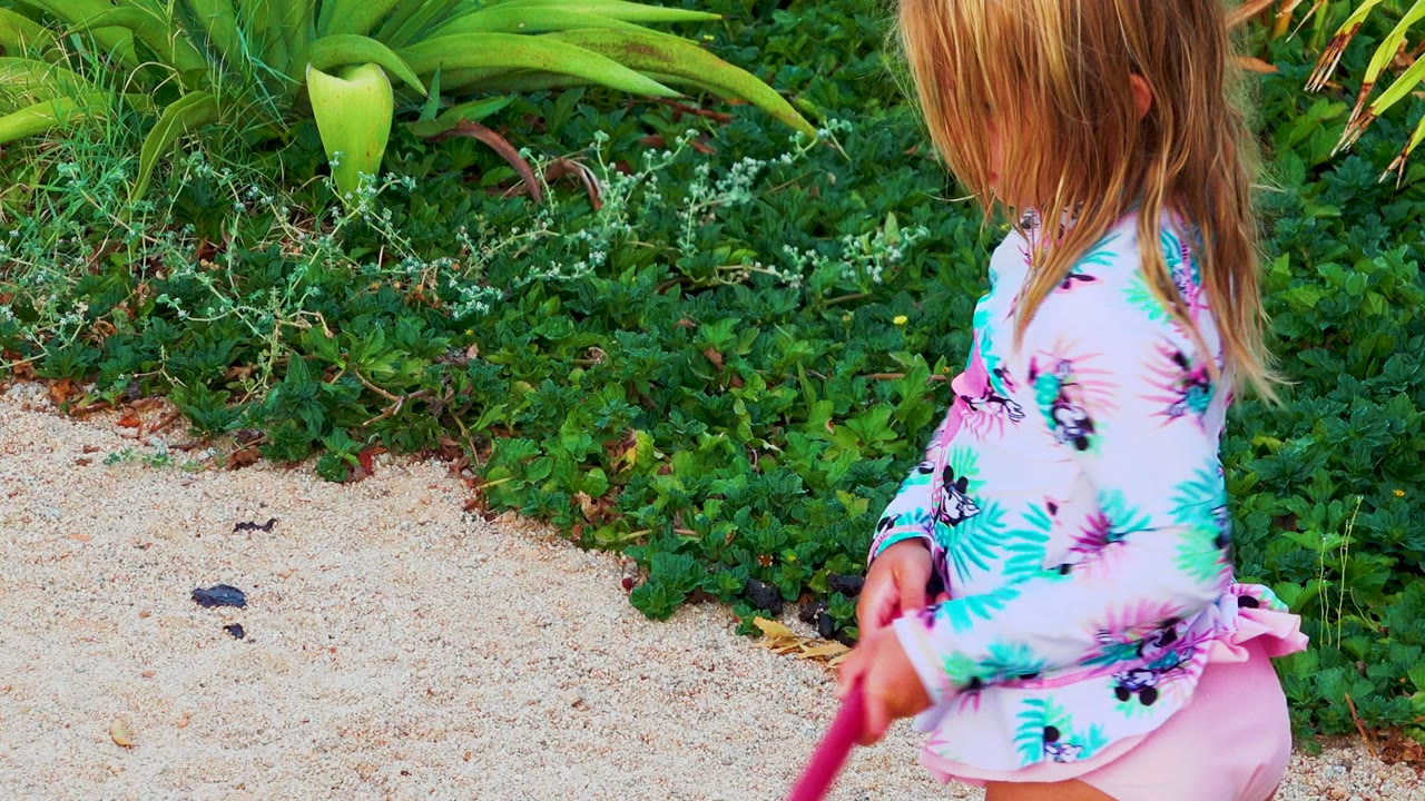 Young girl partly in frame with a red shovel near sand and foliage.