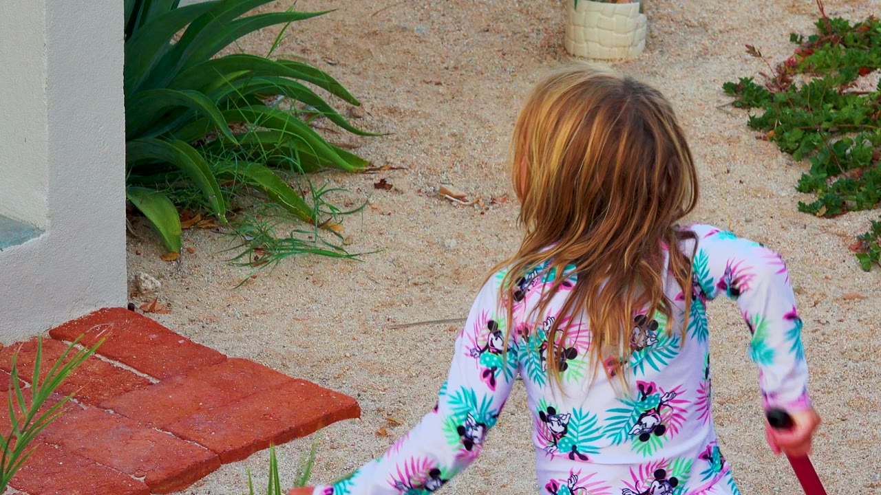 Young blonde girl in a pink patterned bathing suit is seen from behind near red pavers, sandy ground, white walls, and green plants in the 2020 El Sargento La Ventana collection.