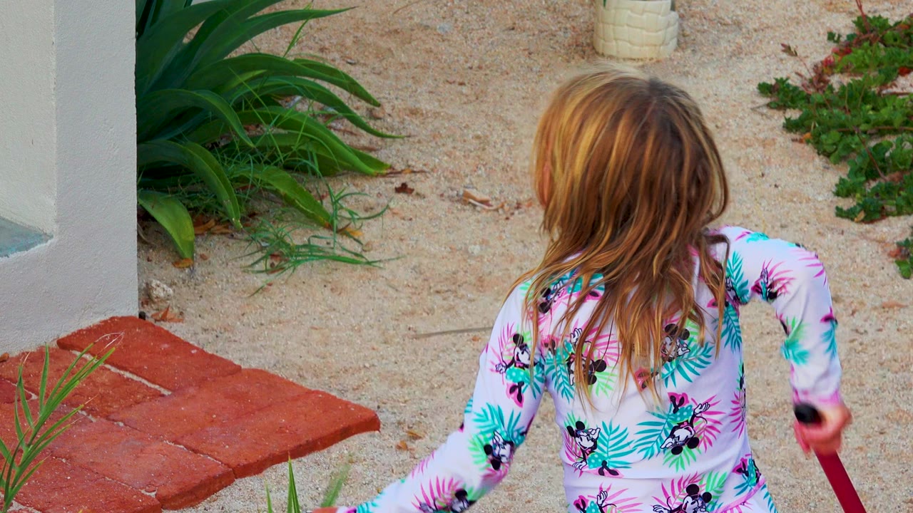 The girl remains turned away with wet-looking blonde hair, holding the pink stick near red pavers and sandy garden planting in the 2020 El Sargento La Ventana collection.