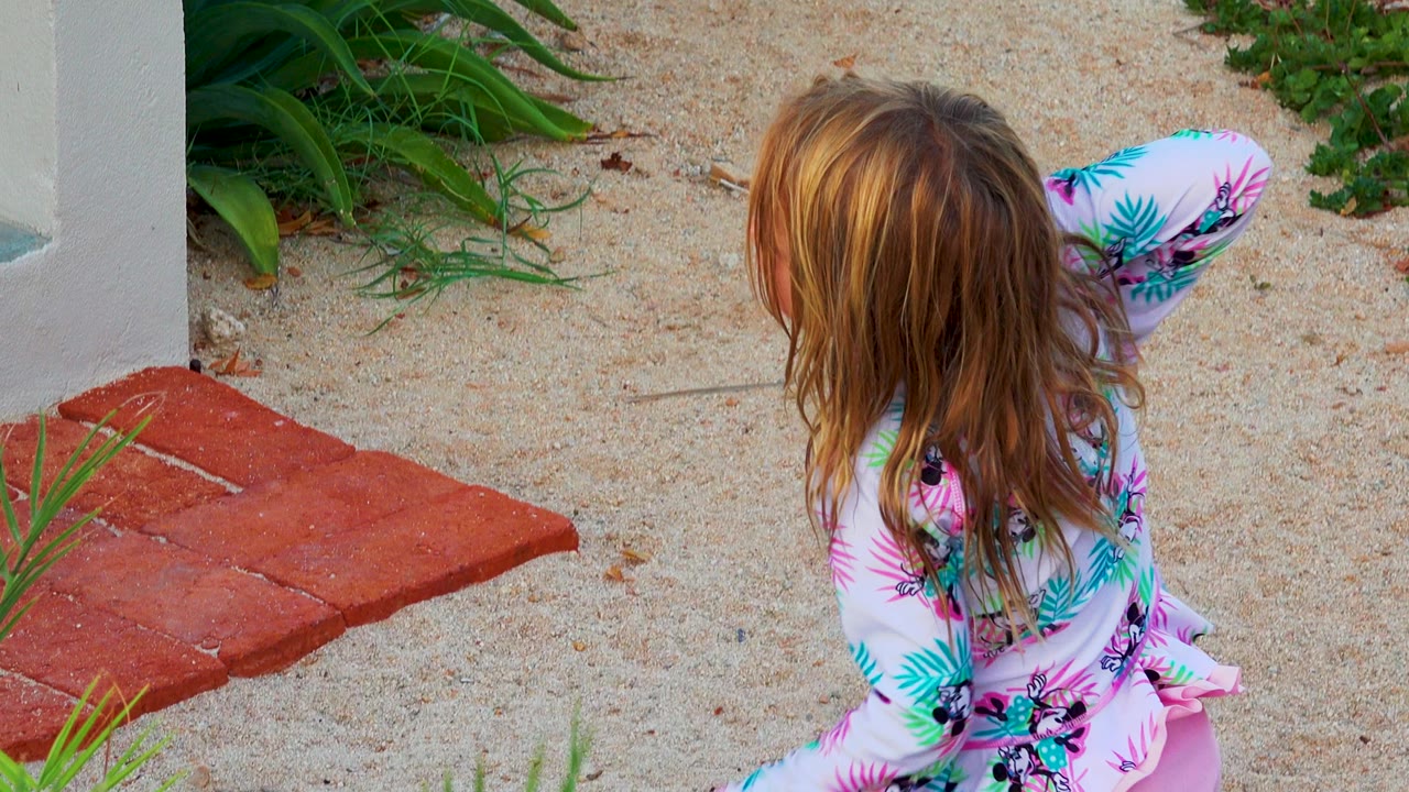 The child turns partly to the side and lifts the pink stick across the sandy garden area, with red pavers, white walls, and green planting around her.