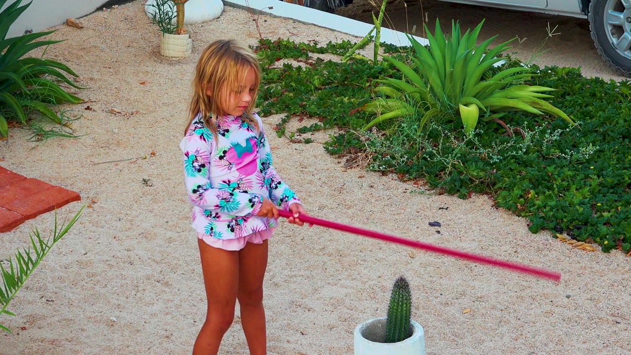 Small blond girl in a pink patterned swim top holds a long pink pole while standing on pale gravel in a Mexico garden with green plants behind her.