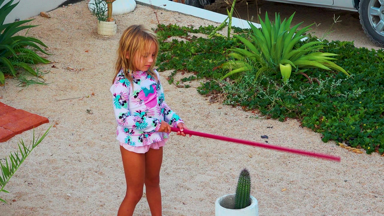 The girl looks down at the pink pole in her hands, with low greenery, agave-like plants, gravel, and a white planter visible around the Mexico garden.