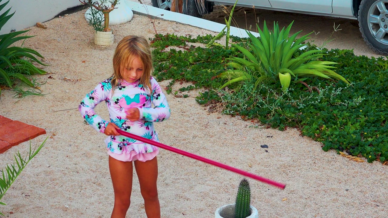 The child faces forward while holding the pink pole diagonally across her body, with pale gravel, green planting beds, and a white planter in the Mexico garden.