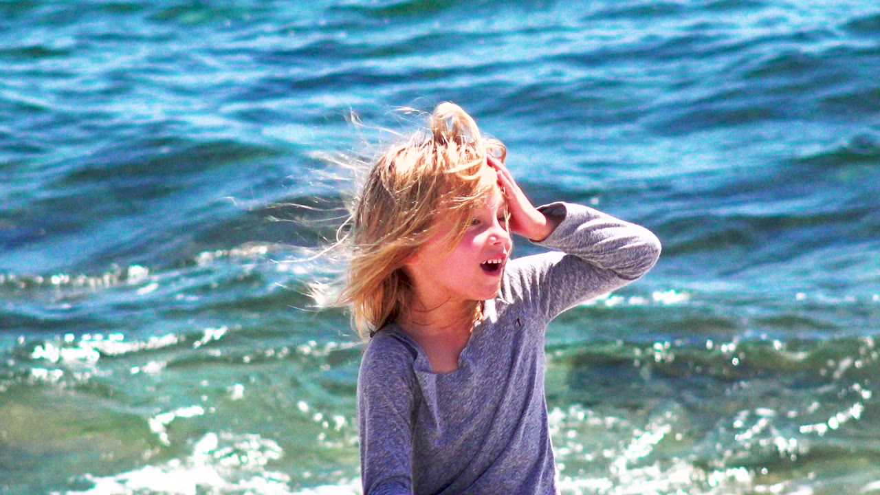 Blond child in a gray long-sleeve shirt sits near the surf in El Sargento La Ventana, Mexico, with one hand raised to the side of the head and blue water behind.