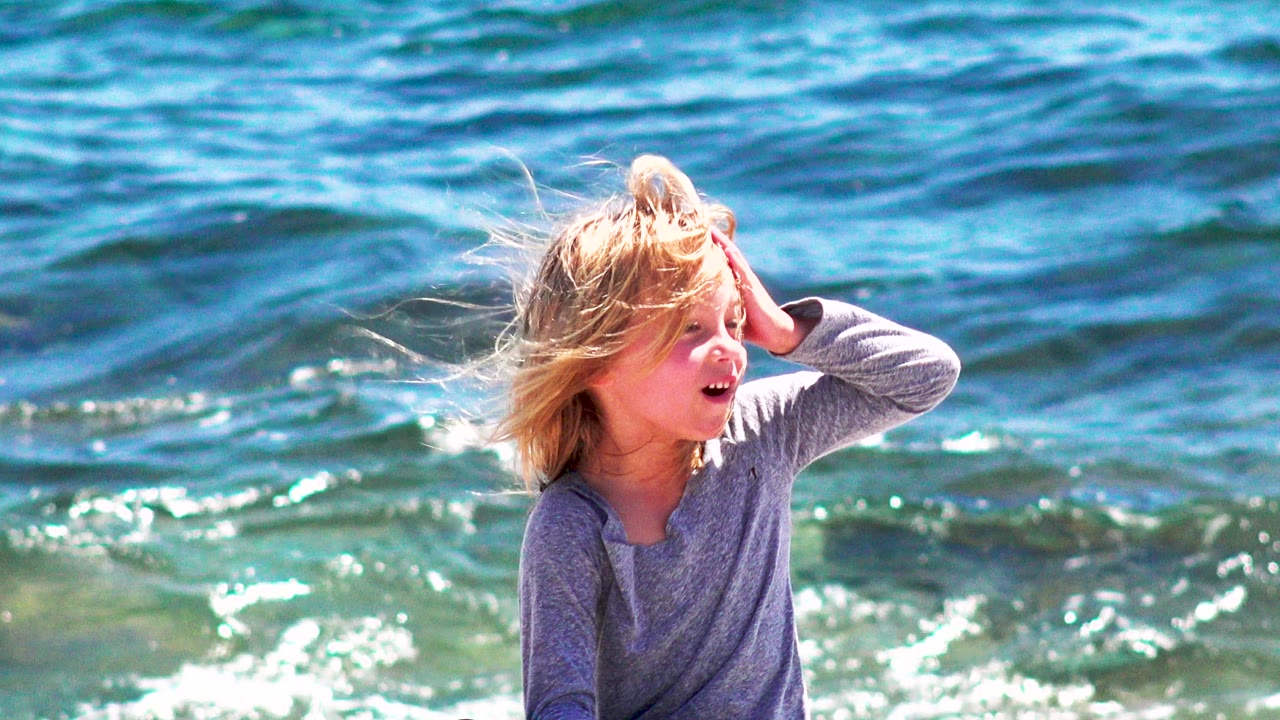 Child near surf with windblown hair and hand at head.