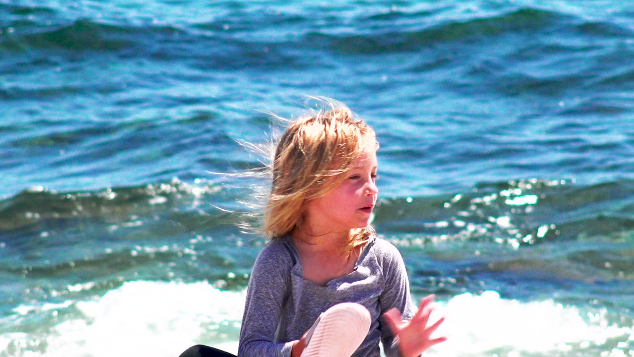 The child turns to the side at the water's edge in El Sargento La Ventana, Mexico, with a white-soled shoe visible and bright foam breaking behind.