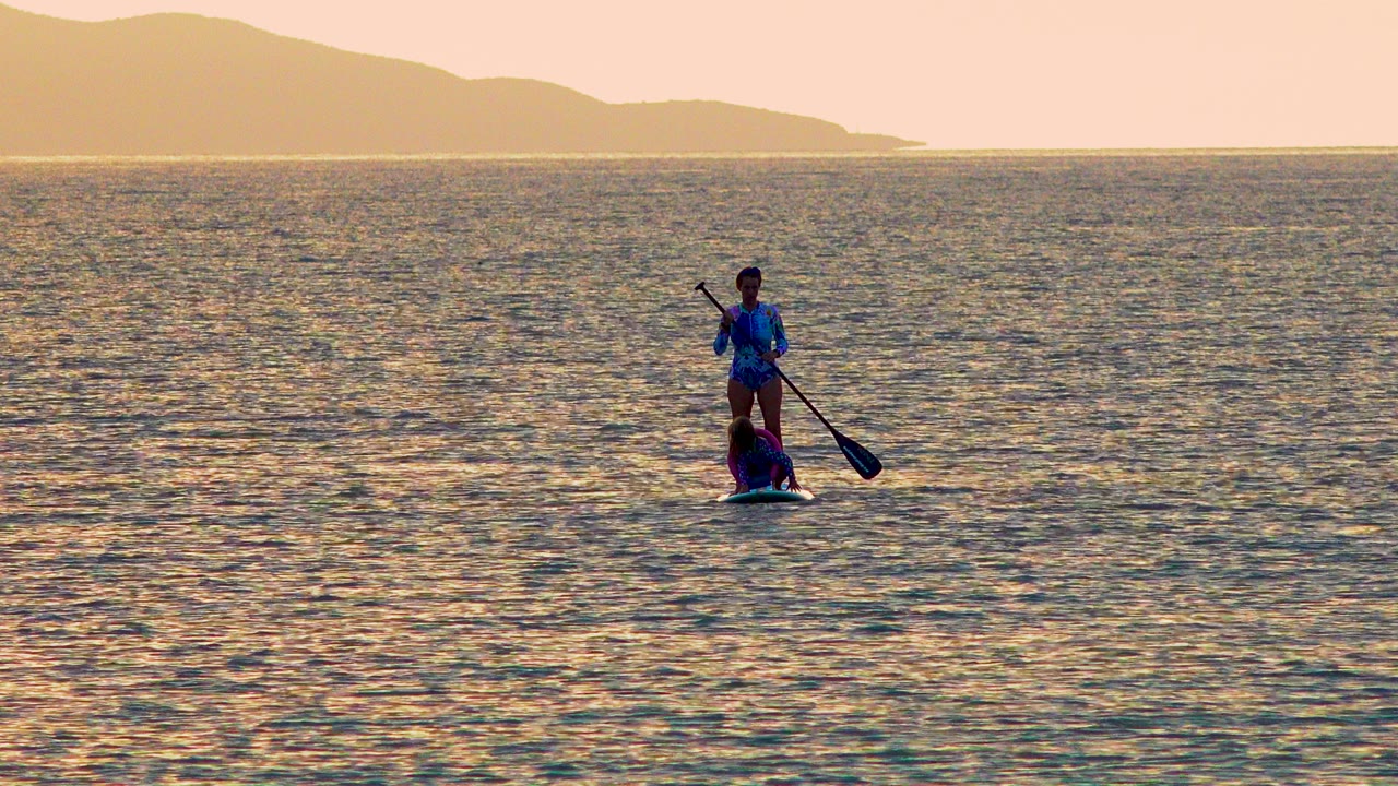 A distant paddleboarder with a seated passenger moves across golden ocean water in El Sargento La Ventana, Mexico, with hazy hills on the horizon.