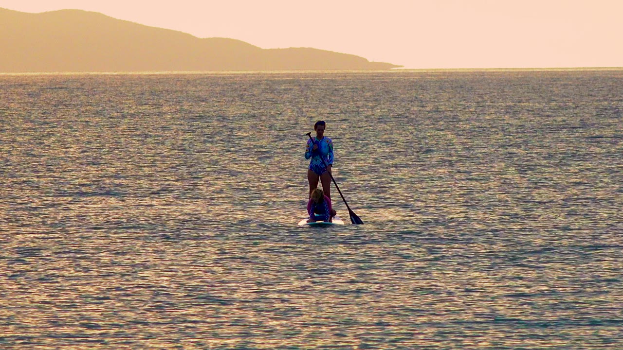 A small stand-up paddleboard carrying two people appears farther right on the warm ocean surface in El Sargento La Ventana, Mexico, below a hazy hill line.