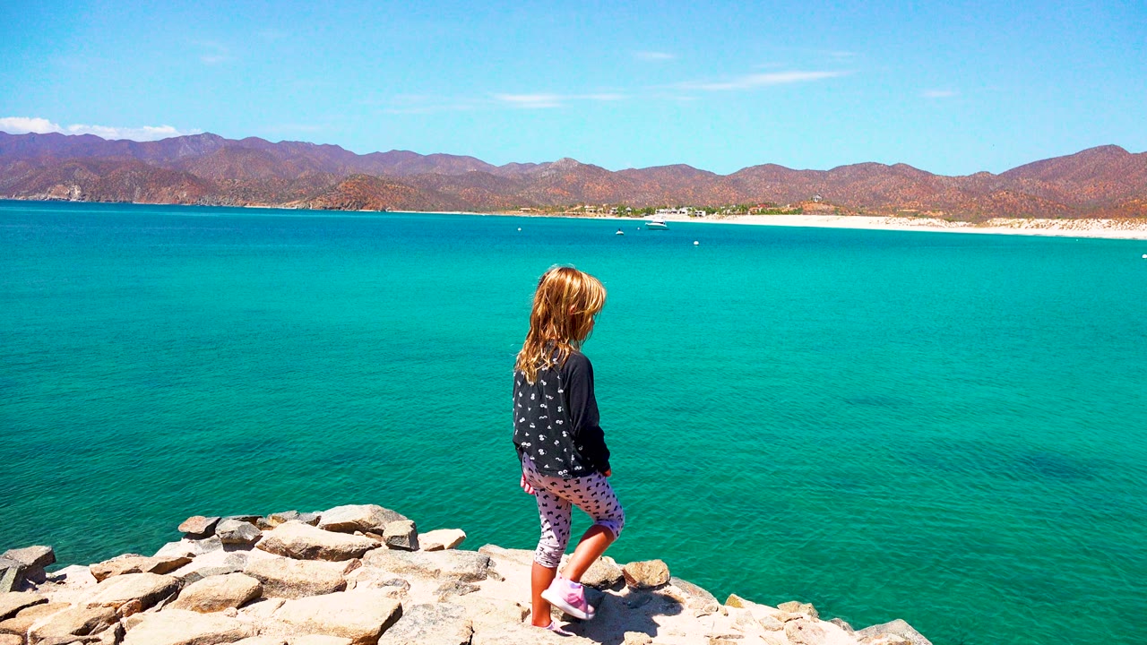 The child is framed near the right edge on coastal rocks, facing the wide turquoise water and low sandy shoreline in El Sargento La Ventana, Mexico.