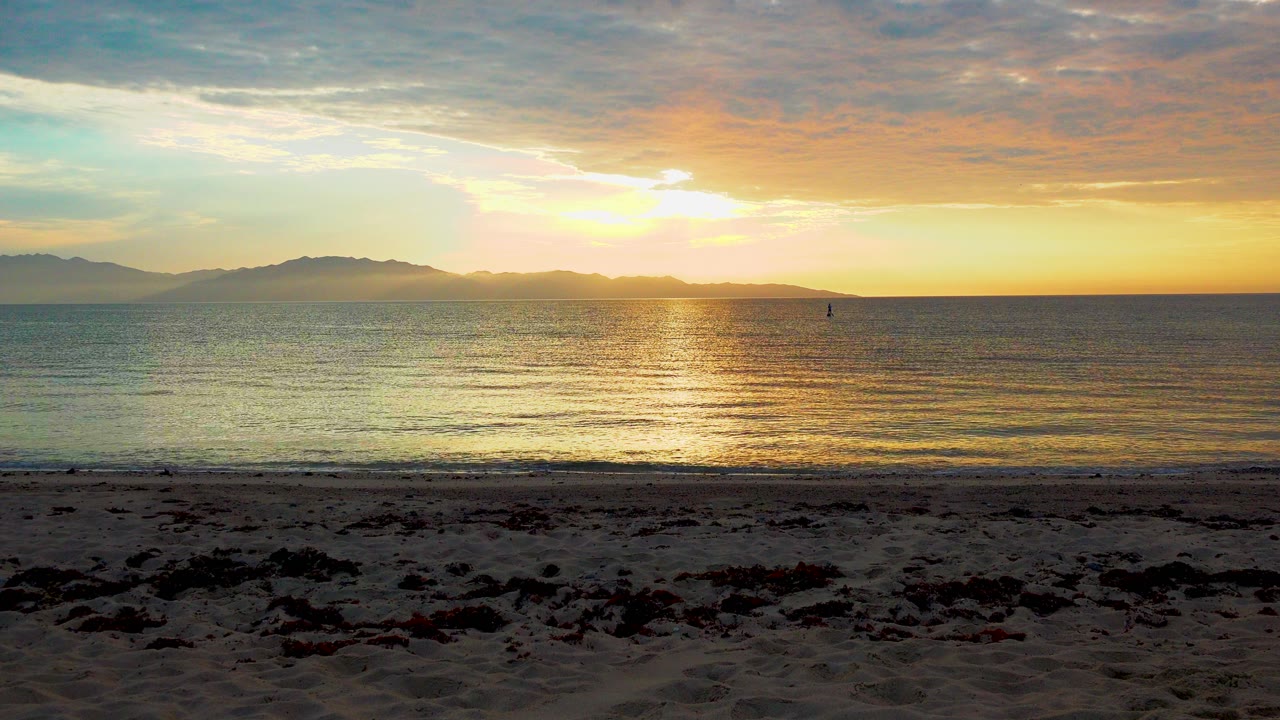 Opening frame of a low sun over the water in El Sargento, Mexico, with a gold reflection, distant mountains, dark seaweed on the sand, and no visible people.