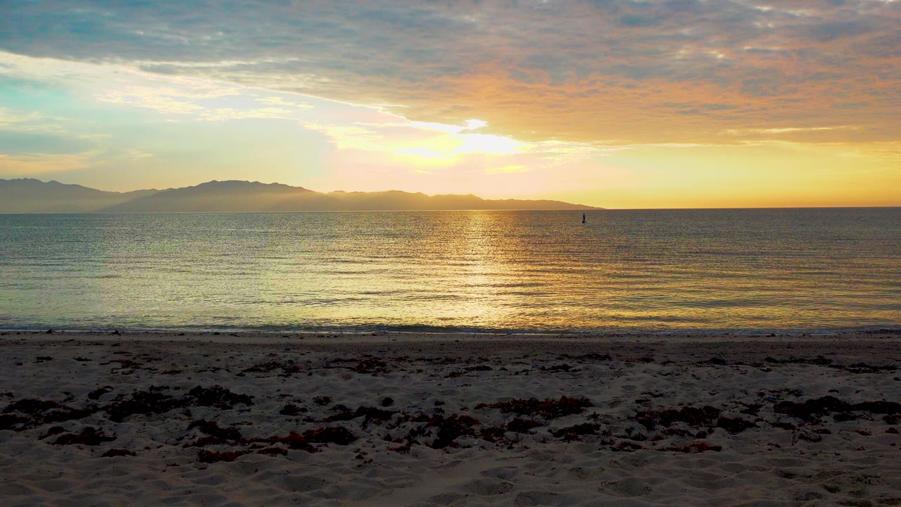 The sun sits just above the horizon in El Sargento, Mexico, sending a bright reflection toward the shore while small waves meet a dark sandy foreground with no visible people.